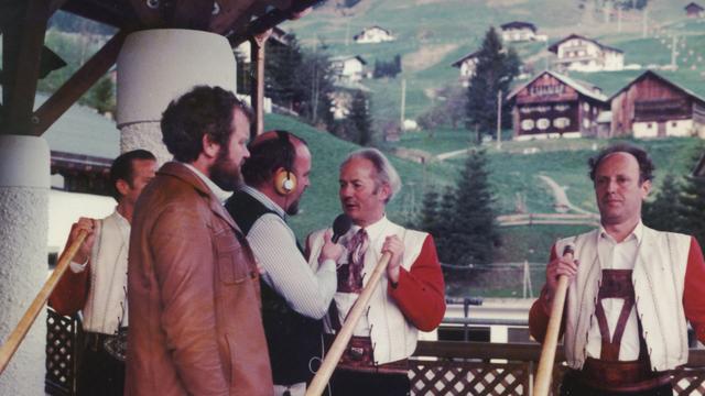 Zwei Reporter interviewen drei Alphornbläser in traditioneller Kleidung vor alpiner Kulisse Zwei Reporter interviewen drei Alphornbläser in traditioneller Kleidung vor alpiner Kulisse