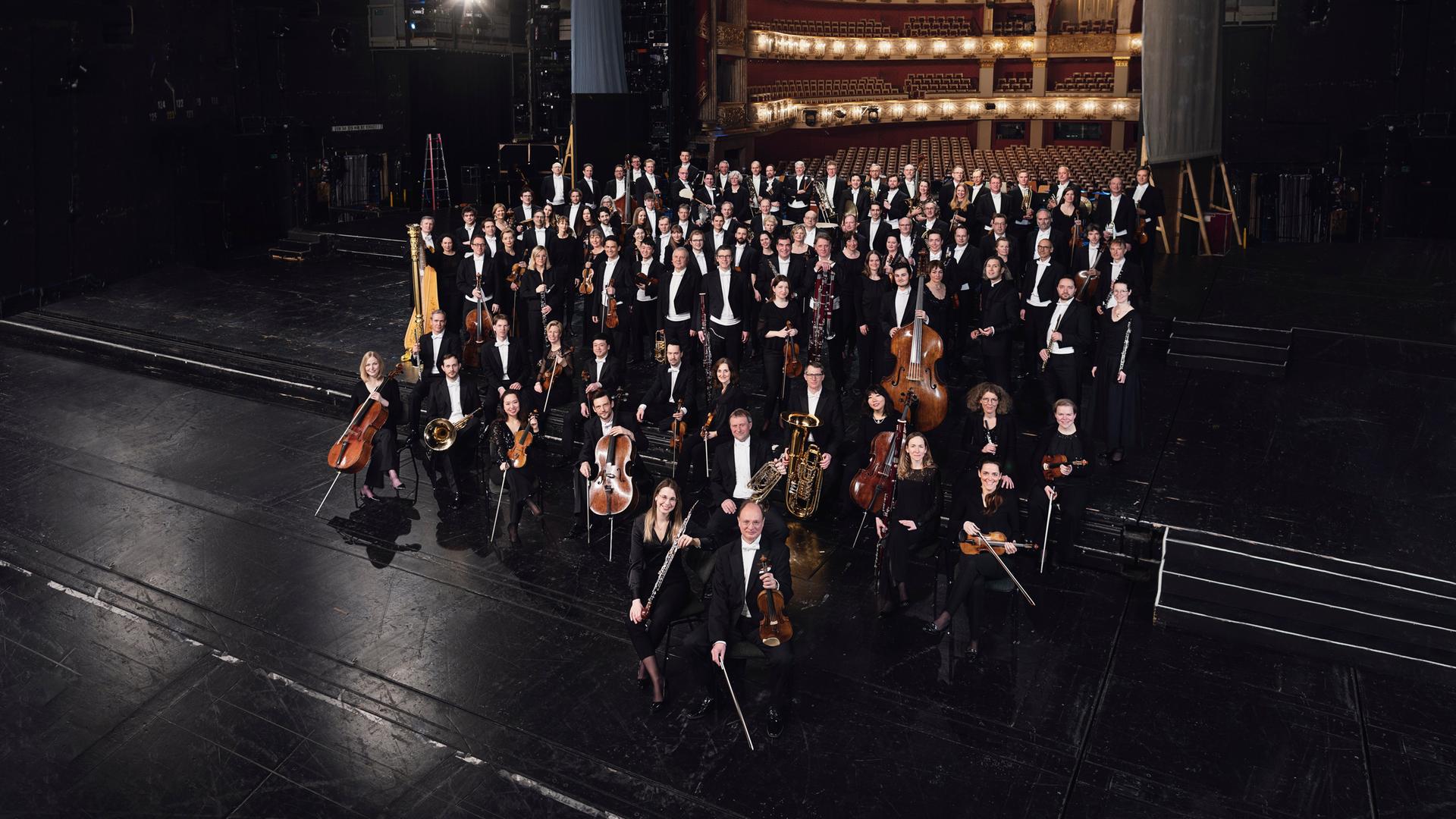 Orchestermitglieder stehen mit ihren Instrumenten auf einer schwarzen Opernbühne. Im Hintergrund ist ein feierlich erleuchteter, leerer Saal zu sehen.