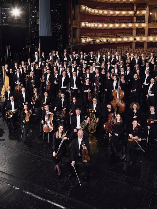 Orchestermitglieder stehen mit ihren Instrumenten auf einer schwarzen Opernbühne. Im Hintergrund ist ein feierlich erleuchteter, leerer Saal zu sehen.