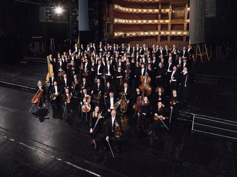 Orchestermitglieder stehen mit ihren Instrumenten auf einer schwarzen Opernbühne. Im Hintergrund ist ein feierlich erleuchteter, leerer Saal zu sehen.