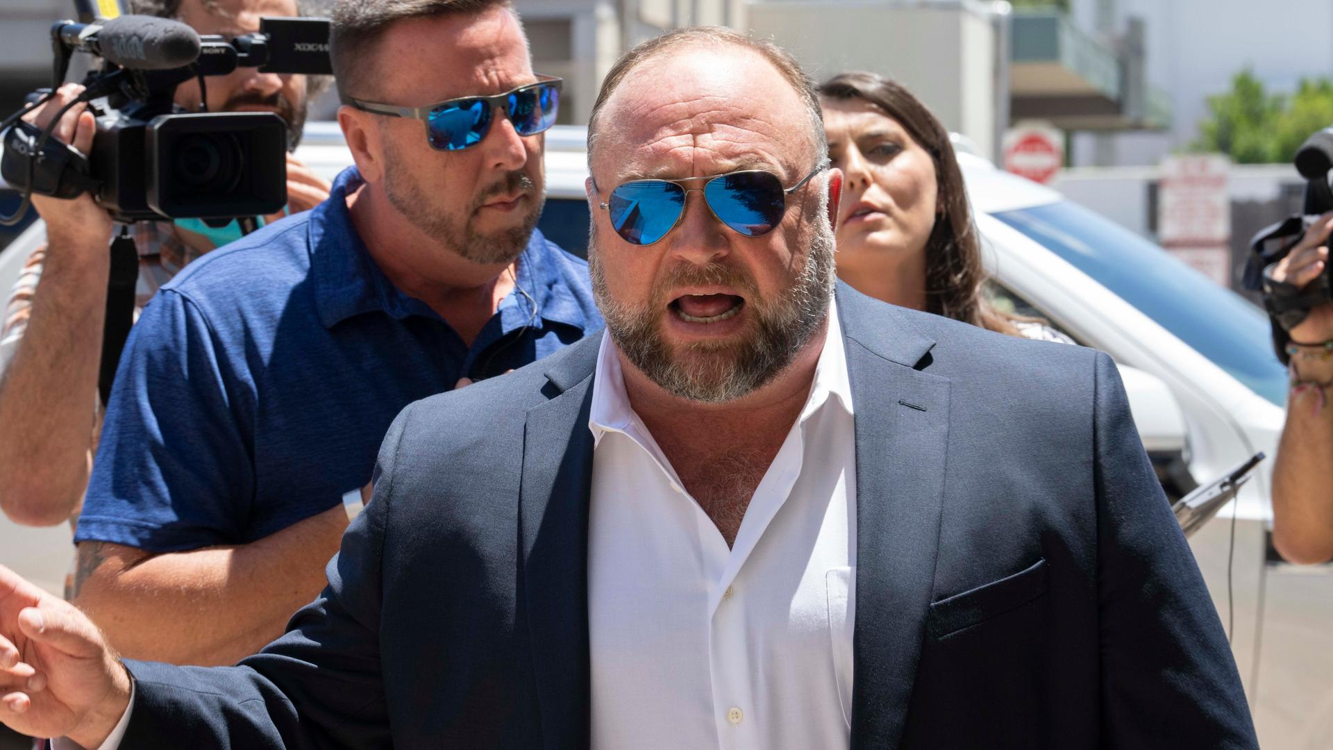Ein korpulenter Mann mit spiegelnder Sonnenbrille steht vor einem Gerichtsgebäude, im Hintergrund sind Medienvertreterinnen und -vertreter zu sehen. Es ist der US-amerikanische Verschwörungsideologe und Verbreiter von Fake News, Alex Jones. Ein korpulenter Mann mit spiegelnder Sonnenbrille steht vor einem Gerichtsgebäude, im Hintergrund sind Medienvertreterinnen und -vertreter zu sehen. Es ist der US-amerikanische Verschwörungsideologe und Verbreiter von Fake News, Alex Jones.
