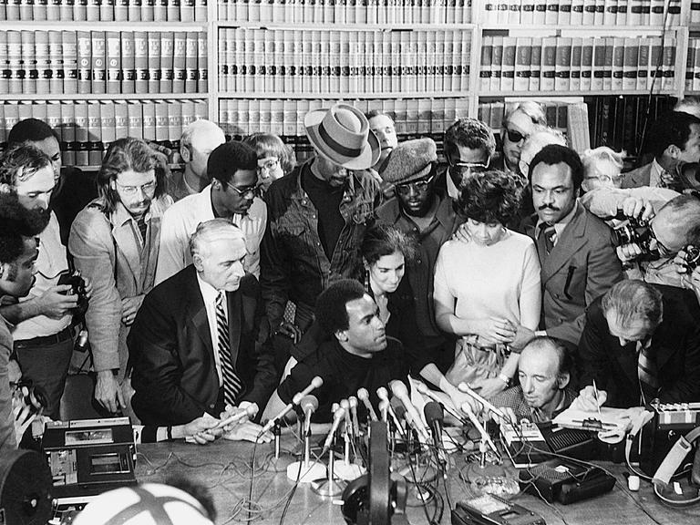 Der US-Aktivist Huey Newton sitzt bei einer Pressekonferenz 1971 an einem Tisch mit einer Reihe von Mikrofonen und wird umringt von einer Gruppe von Menschen