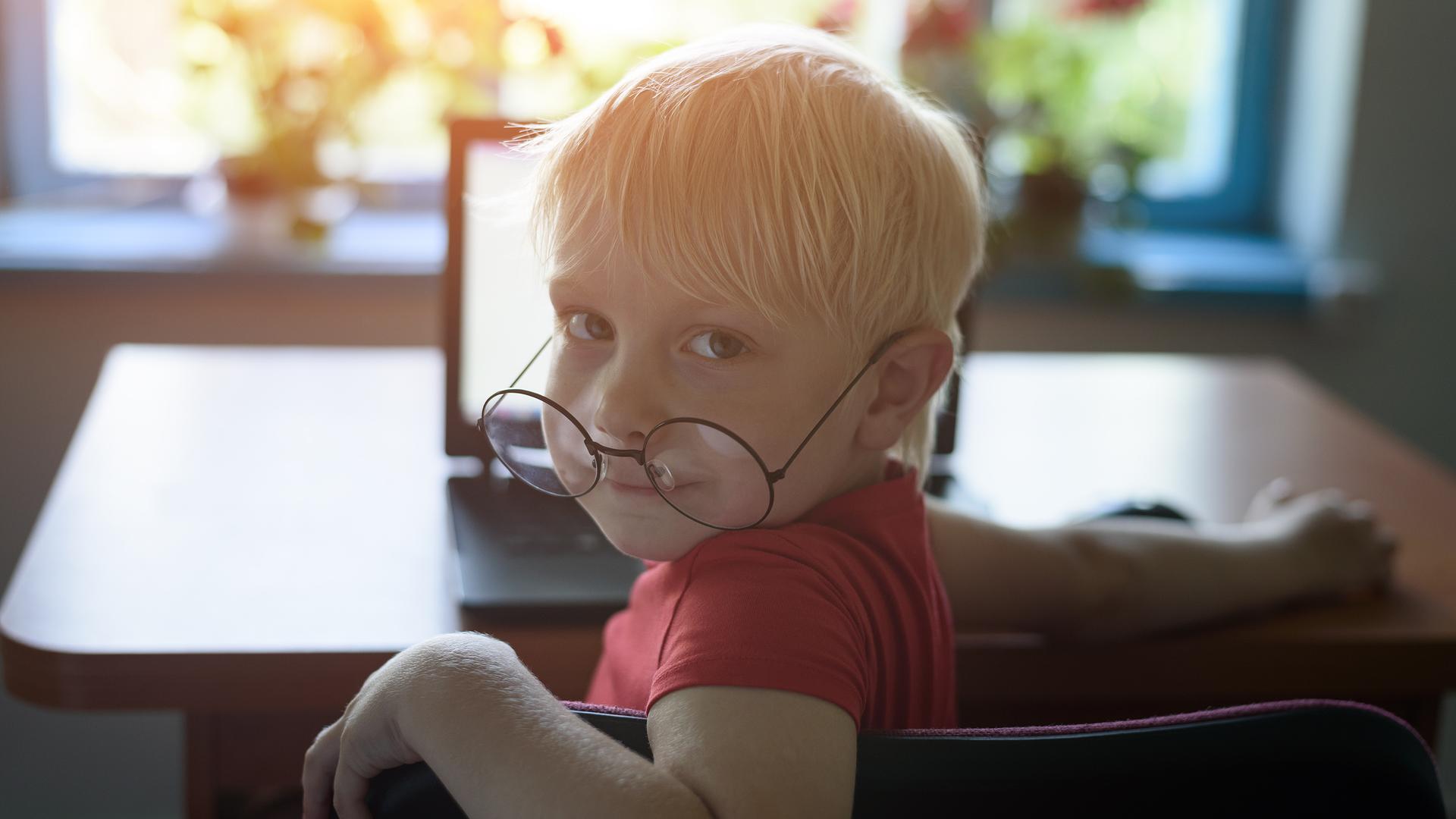 Ein Junge mit einer übergroßen Brille auf der Nase sitzt vor einem Laptop und blickt über die Schulter in die Kamera. Ein Junge mit einer übergroßen Brille auf der Nase sitzt vor einem Laptop und blickt über die Schulter in die Kamera.