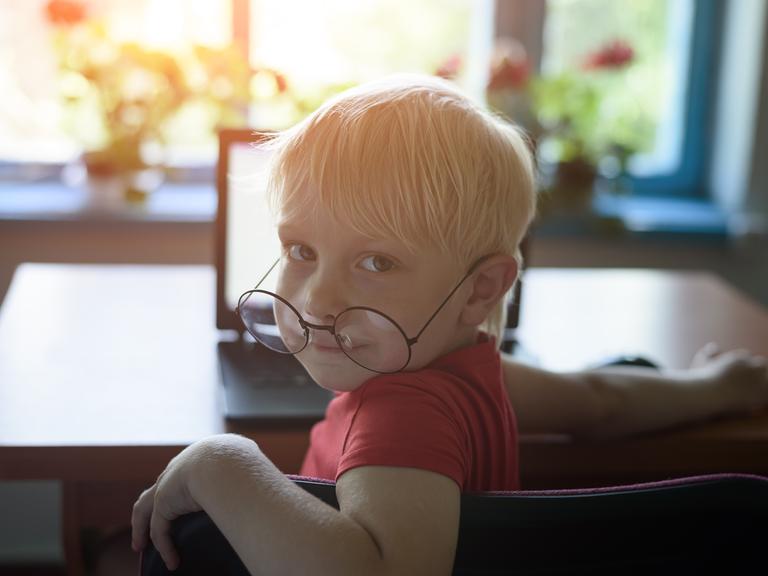 Ein Junge mit einer übergroßen Brille auf der Nase sitzt vor einem Laptop und blickt über die Schulter in die Kamera.