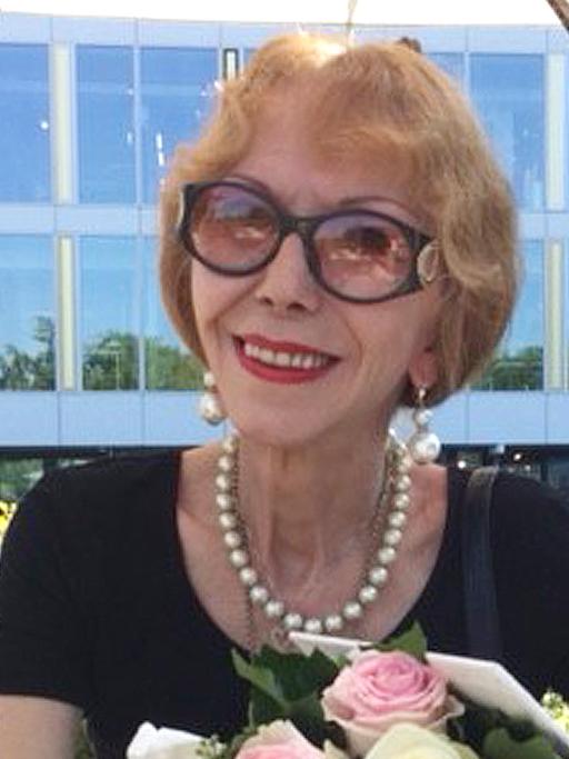 Rosemarie Schubert-Blum. Eine ältere Frau mit halblangen Haaren und Sonnenbrille sitzt auf der Terrasse eines Kaffees. 