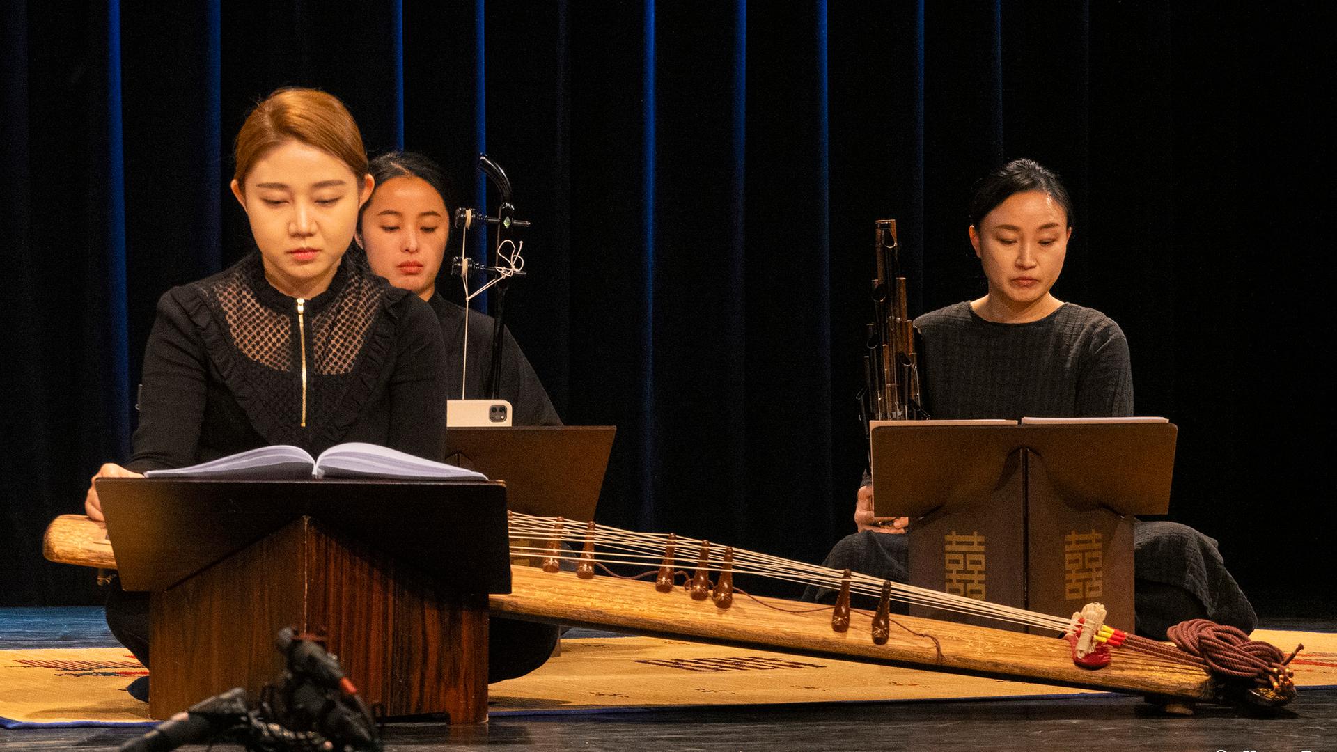 Drei Musikerinnen mit traditionellen koreanischen Instrumenten sitzen mit kontrollierten Blicken auf der Bühne