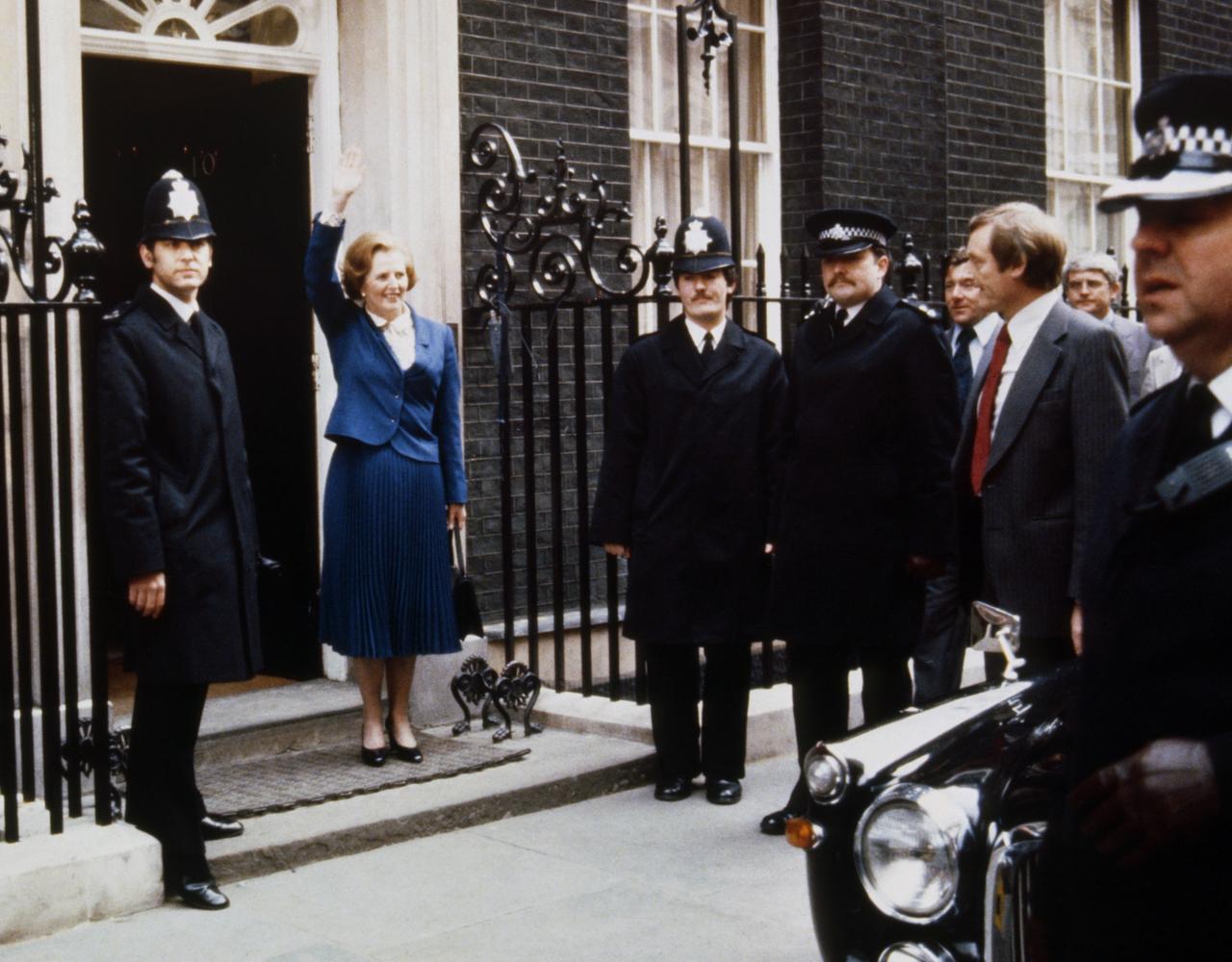 Margaret Thatcher steht, umringt von Polizisten, am Eingang der Downing Street Nr. 10 und winkt Personen die im Bild nicht zu sehen sind.