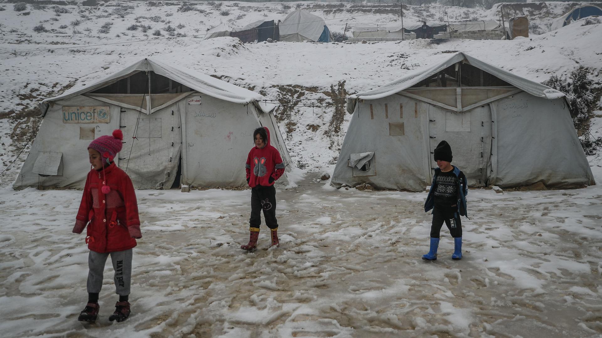 Drei Kinder stehen vor Zelten in einem Flüchtlingslager. Es liegt Schnee. Drei Kinder stehen vor Zelten in einem Flüchtlingslager. Es liegt Schnee.