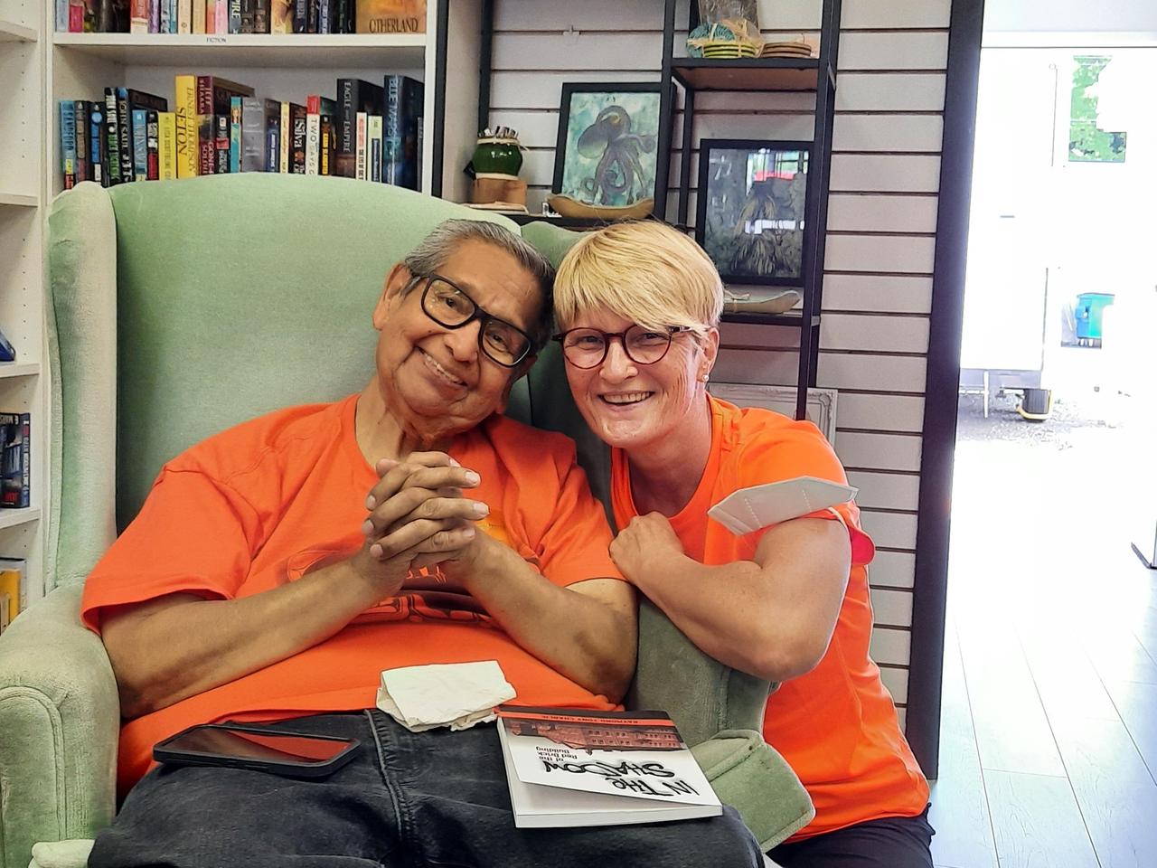 Ein alter Mann sitzt in einem Sessel und lehnt sich an eine blonde Frau an, beide tragen orangene T-Shirts und lachen in die Kamera. Es sind Raymond Tony Charlie mit der Autorin im Buchlanden von Warren Goulding in Chemainus