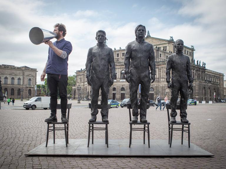 Edward Snowden, Julian Assange und Chelsea Manning als Skulpturen der Kunstaktion "Anything to say" im Jahr 2015 stehen auf drei Stühlen  auf dem Theaterplatz in Dresden, links daneben steht der Künstler Davide Domino.