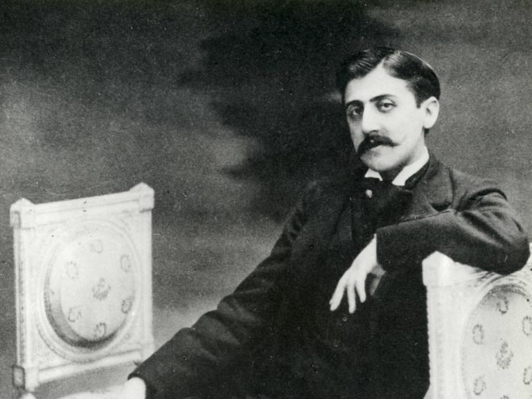 Auf einer alten Fotografie sitzt der Schriftsteller Marcel Proust,leger auf einer Lounge und blickt in die Kamera.