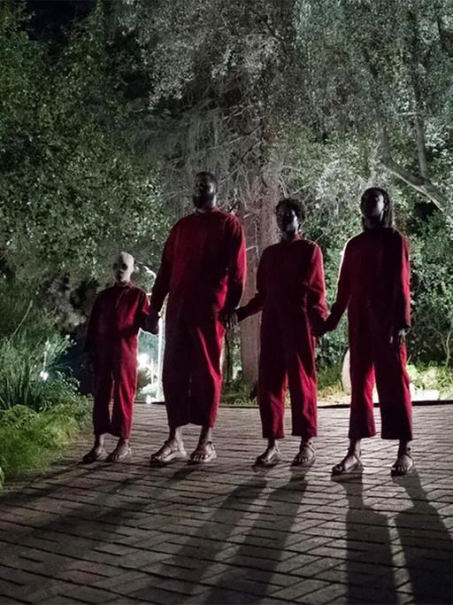 Vier Personen in roten Jumpsuits stehen Hand in Hand bei nächtlicher Beleuchtung vor einem Haus. (Szenenbild aus dem Film "Wir" von Jordan Peele.)