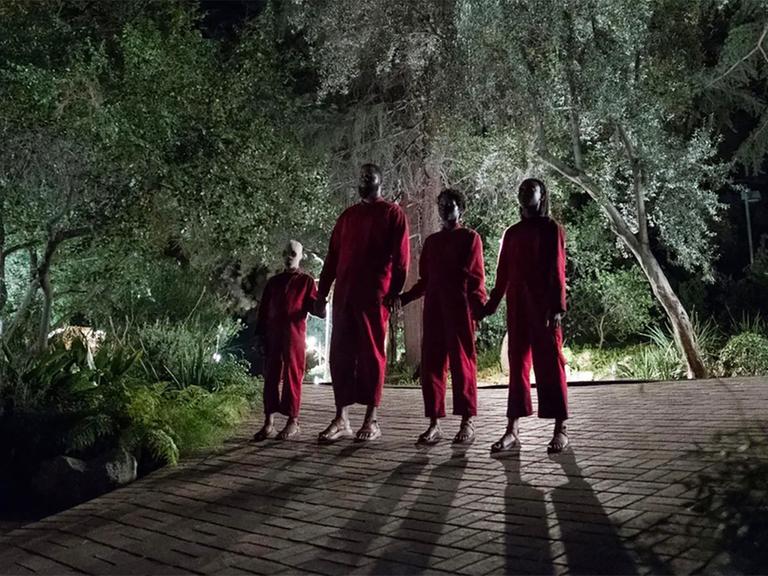 Vier Personen in roten Jumpsuits stehen Hand in Hand bei nächtlicher Beleuchtung vor einem Haus. (Szenenbild aus dem Film "Wir" von Jordan Peele.)