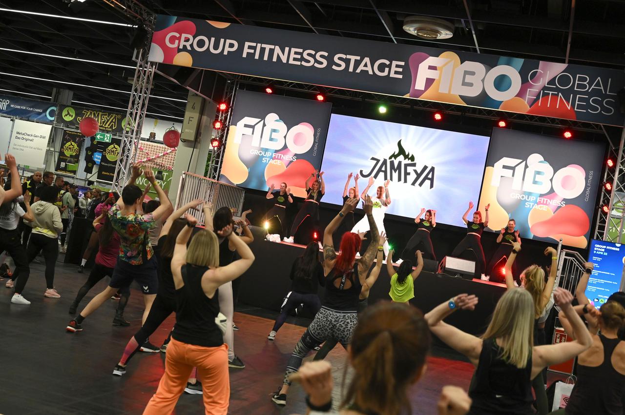 Menschen nehmen vor einer Bühne an einem Gymnastikkurs teil. Die Bühne ist überschrieben mit den Worten: "Group Fitness Stage. FIBO Global Fitness".