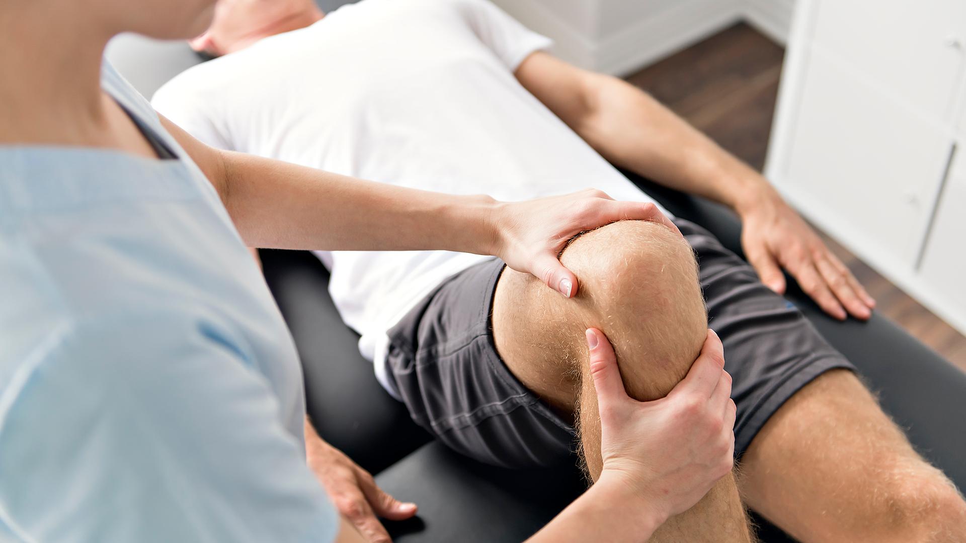Ein Patient liegt auf einem Behandlungstisch und wird von einer Physiotherapeutin am Knie behandelt. Ein Patient liegt auf einem Behandlungstisch und wird von einer Physiotherapeutin am Knie behandelt.