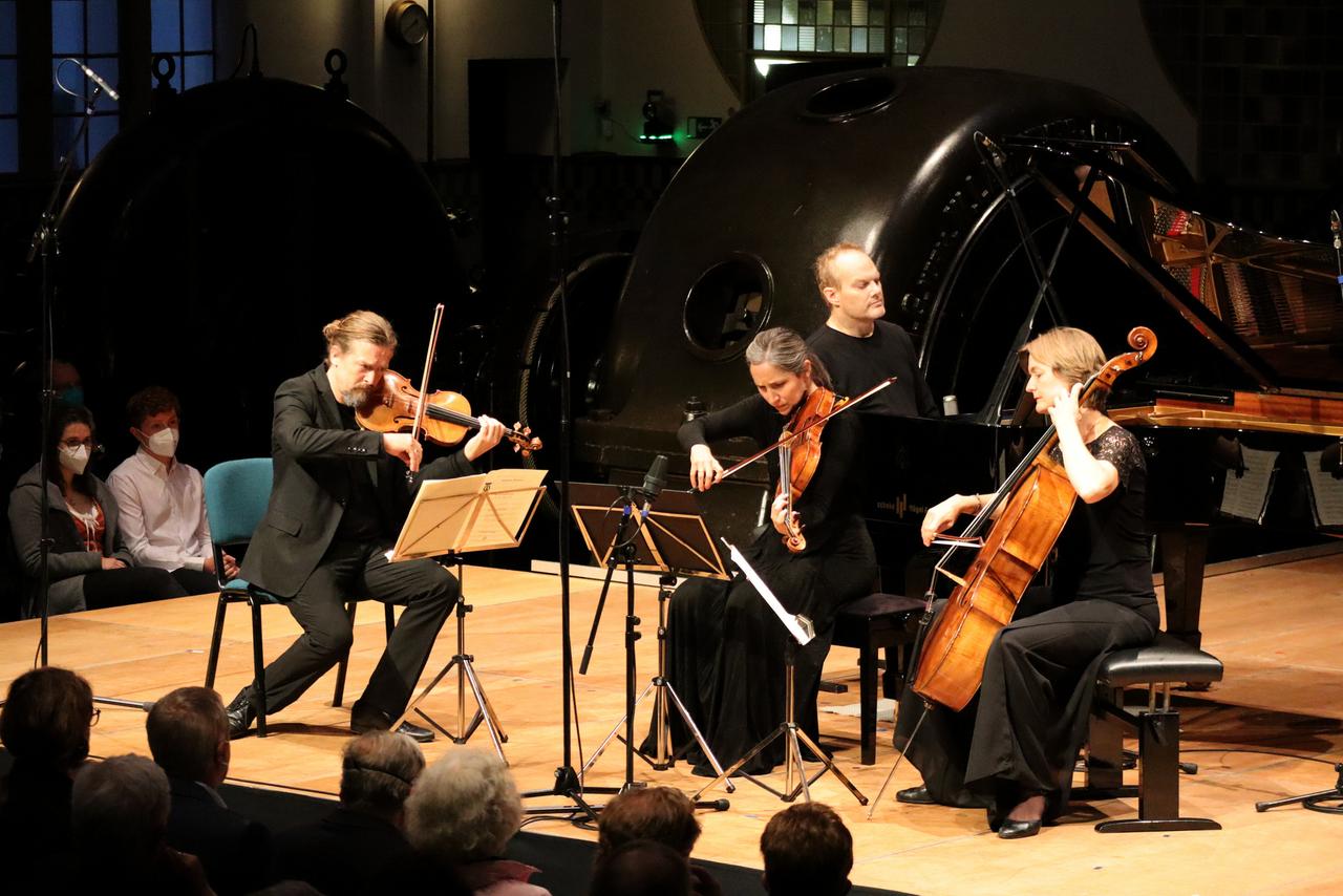 Ein Geiger, eine Bratschistin, ein Pianist und eine Cellistin musizieren auf einer Bühne. Im Hintergrund sind Maschinen des Jugendstil-Kraftwerks Heimbach (Eifel) sowie einige Zuhörerinnen und Zuhörer zu sehen.