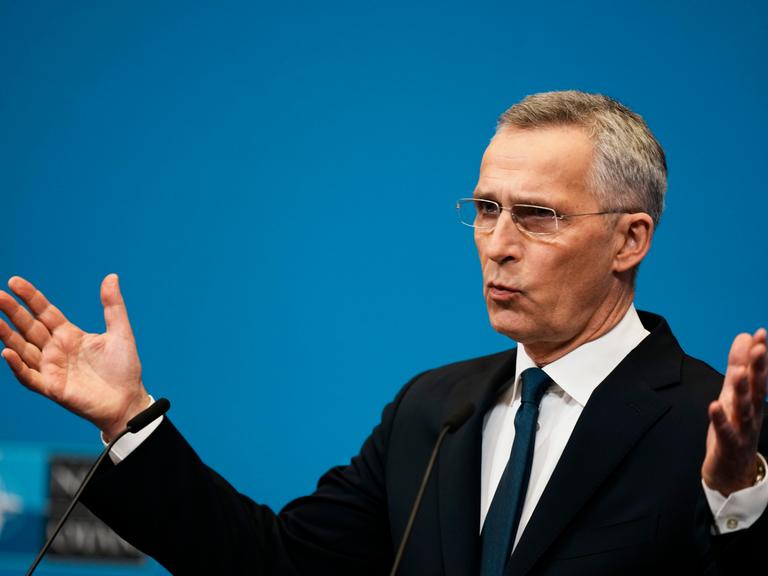 Ein Mann in dunklem Anzug hält die Arme ausgebreitet und spricht vor einem blauen Hintergrund.