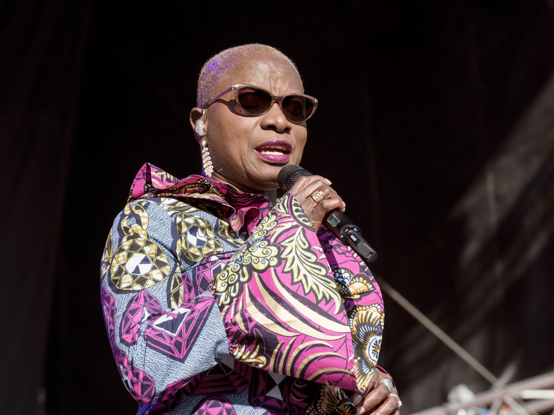 Eine Frau steht auf der Bühne und singt. Sie trägt Sonnenbrille und ein farbenfrohes Kleid. 