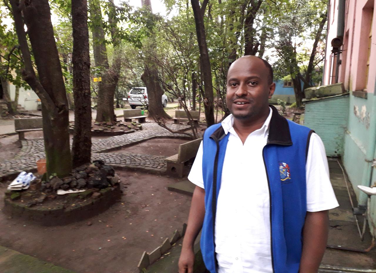 Der äthiopische Pharmakologe Solomon Tesfaye trägt ein weißes Polohemd und eine blaue Weste. Er vor einem Gebäude und lacht in die Kamera.