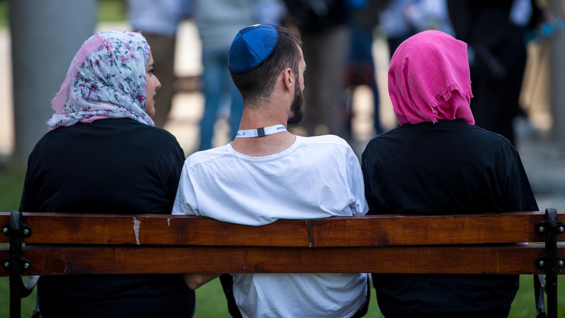 Ein jüdischer Mann mit Kippa sitzt zwischen zwei muslimischen Frauen auf einer Bank. Beide Frauen tragen Kopftuch. Ein jüdischer Mann mit Kippa sitzt zwischen zwei muslimischen Frauen auf einer Bank. Beide Frauen tragen Kopftuch.