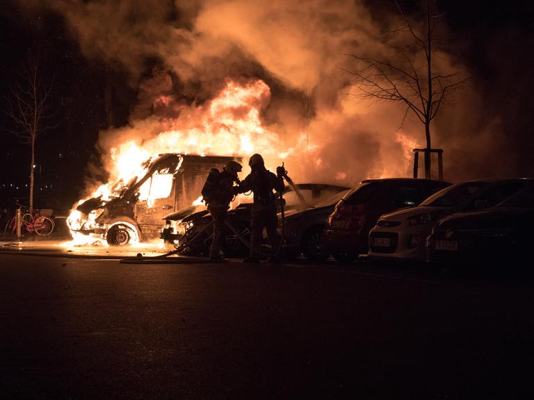 Feuerwehrleute versuchen ein brennendes Auto zu löschen. Es ist Nacht und alles ist sehr dunkel, nur das Feuer sticht hervor.