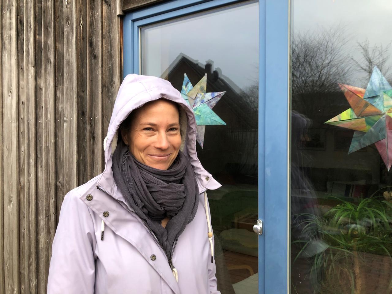 Isabell Wäß steht mit Kapuze auf dem Kopf vor einer Scheibe mit Weihnachtsdekoration an ihrem Holzhaus und lächelt in die Kamera.