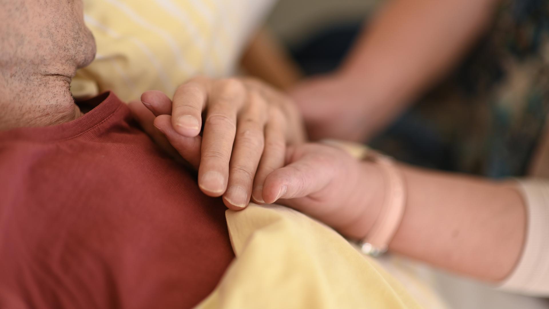 Eine Hospizmitarbeiterin hält die Hand eines todkranken Menschen, der im Hospiz im Bett liegt. Eine Hospizmitarbeiterin hält die Hand eines todkranken Menschen, der im Hospiz im Bett liegt.