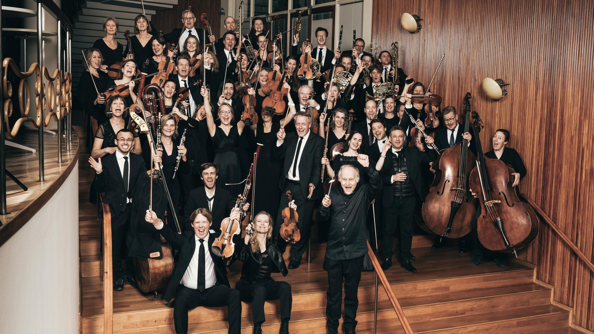 Ein Orchester aus rund 40 Personen steht auf einer Treppe gruppiert. Die Musiker und Musikerinnen halten ihre Instrumente in den Händen und scheinen etwas freudig zu rufen.