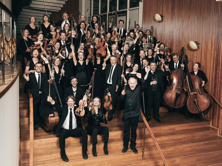 Ein Orchester aus rund 40 Personen steht auf einer Treppe gruppiert. Die Musiker und Musikerinnen halten ihre Instrumente in den Händen und scheinen etwas freudig zu rufen.