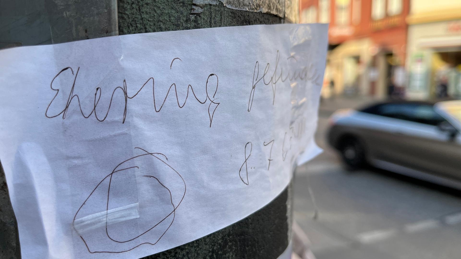 Ein Zettel klebt an einem Ampelmast. Im Hintergrund ist ein Auto auf der Straße erkennbar. Ein Zettel klebt an einem Ampelmast. Im Hintergrund ist ein Auto auf der Straße erkennbar.