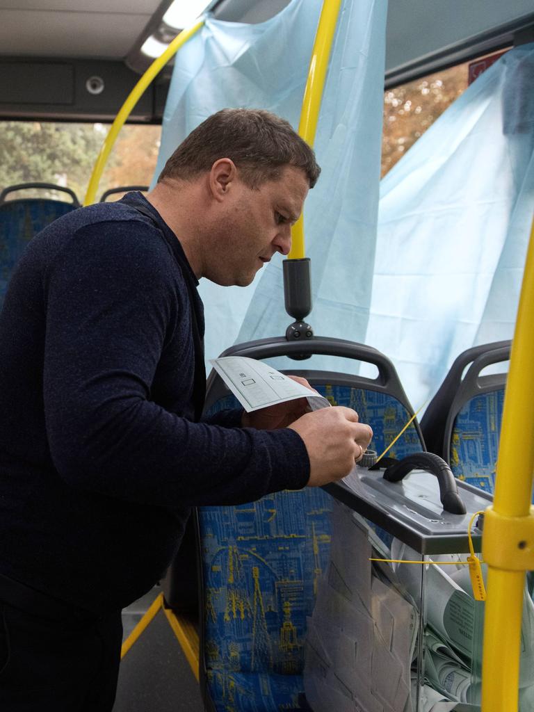 Blick in einen Bus: Ein Polizist sitzt neben einem Mann, der einen bereitgestellten Wahlzettel in eine Plastikschachtel schiebt. 