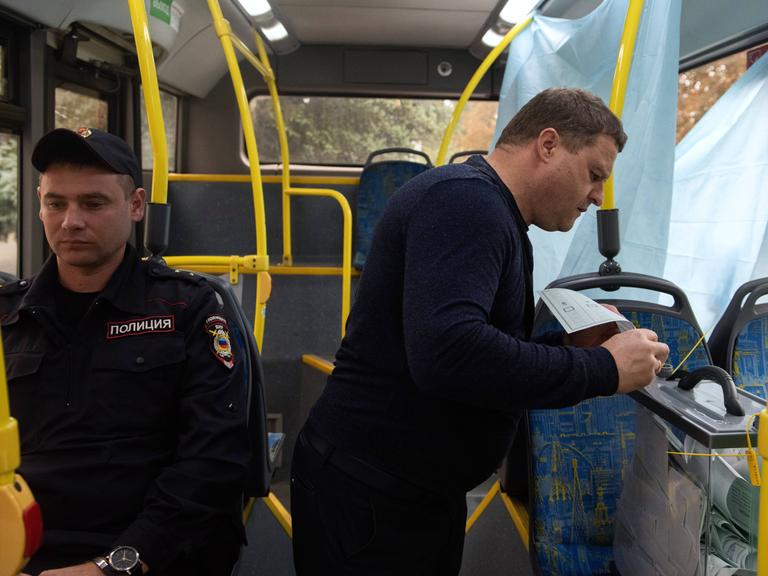 Blick in einen Bus: Ein Polizist sitzt neben einem Mann, der einen bereitgestellten Wahlzettel in eine Plastikschachtel schiebt. 