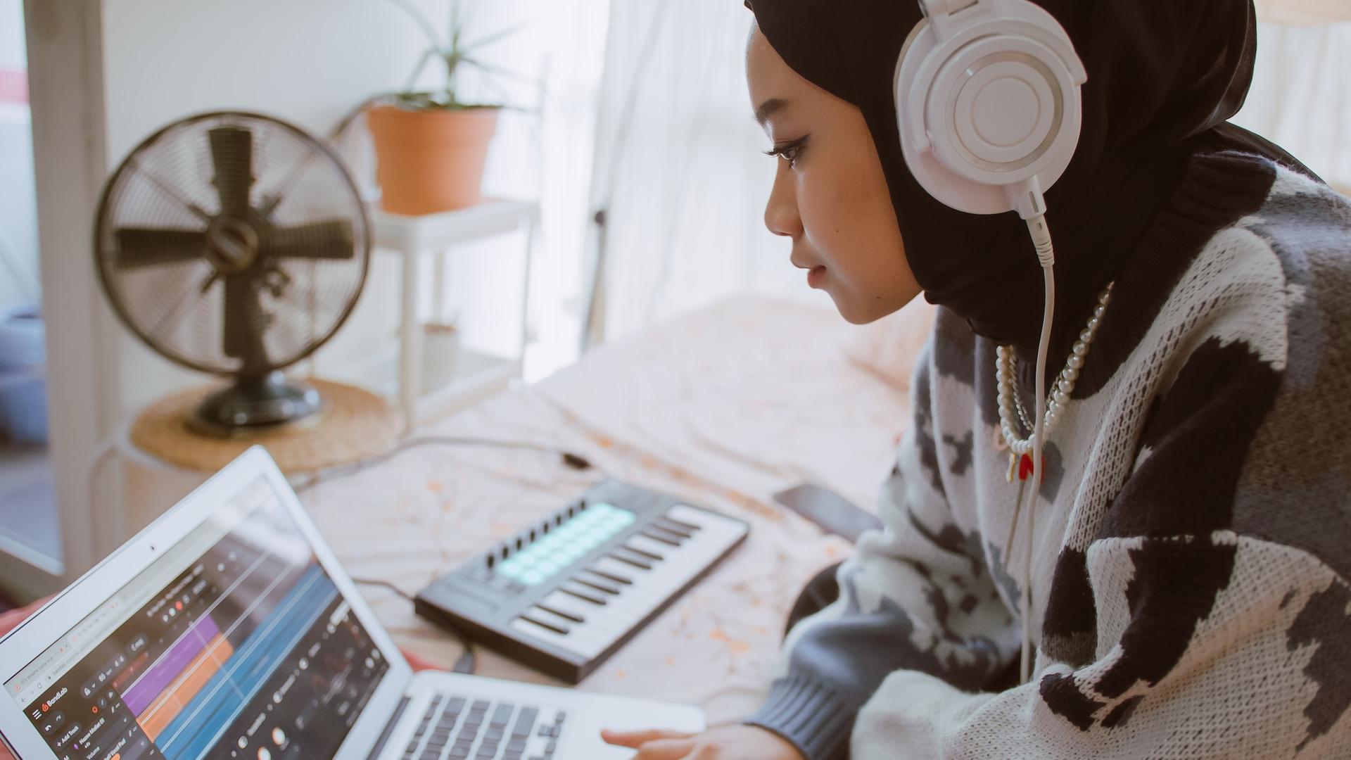 Eine junge, muslimische Frau kreiert Musik auf ihrem Laptop mit Hilfe von Kopfhörern und einer kleinen Piano-Tastatur. Eine junge, muslimische Frau kreiert Musik auf ihrem Laptop mit Hilfe von Kopfhörern und einer kleinen Piano-Tastatur.