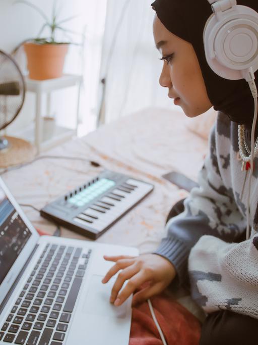 Eine junge, muslimische Frau kreiert Musik auf ihrem Laptop mit Hilfe von Kopfhörern und einer kleinen Piano-Tastatur.