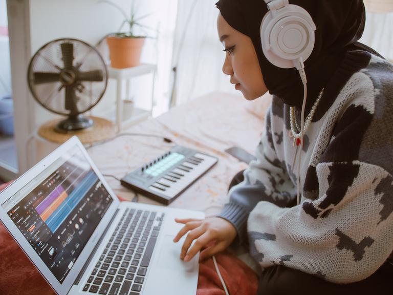 Eine junge, muslimische Frau kreiert Musik auf ihrem Laptop mit Hilfe von Kopfhörern und einer kleinen Piano-Tastatur.