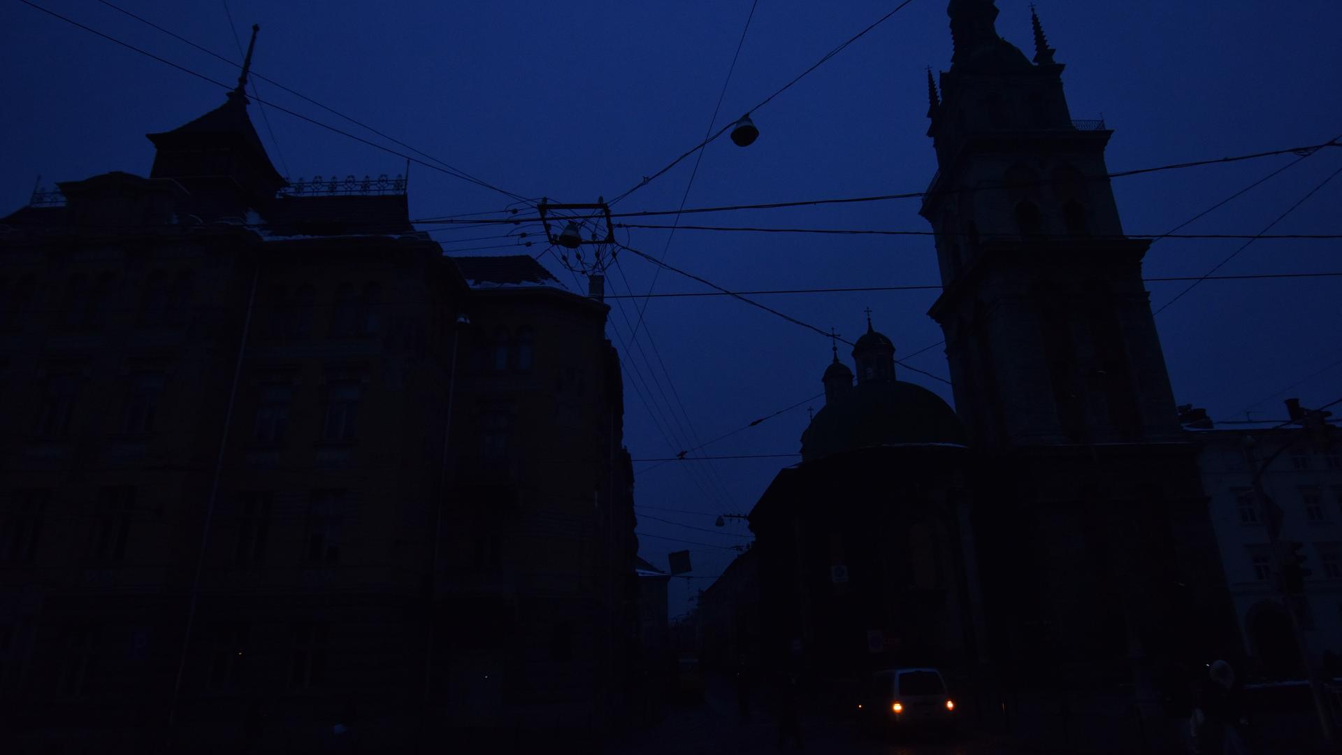 Eine Stadtlandschaft in fast kompletter Dunkelheit. Nur an einer Stelle sind zwei Lichter zu sehen, sonst zeichnen sich lediglich dunkle Gebäudesilhouetten gegen den dunkelblauen Himmel ab. Eine Stadtlandschaft in fast kompletter Dunkelheit. Nur an einer Stelle sind zwei Lichter zu sehen, sonst zeichnen sich lediglich dunkle Gebäudesilhouetten gegen den dunkelblauen Himmel ab.