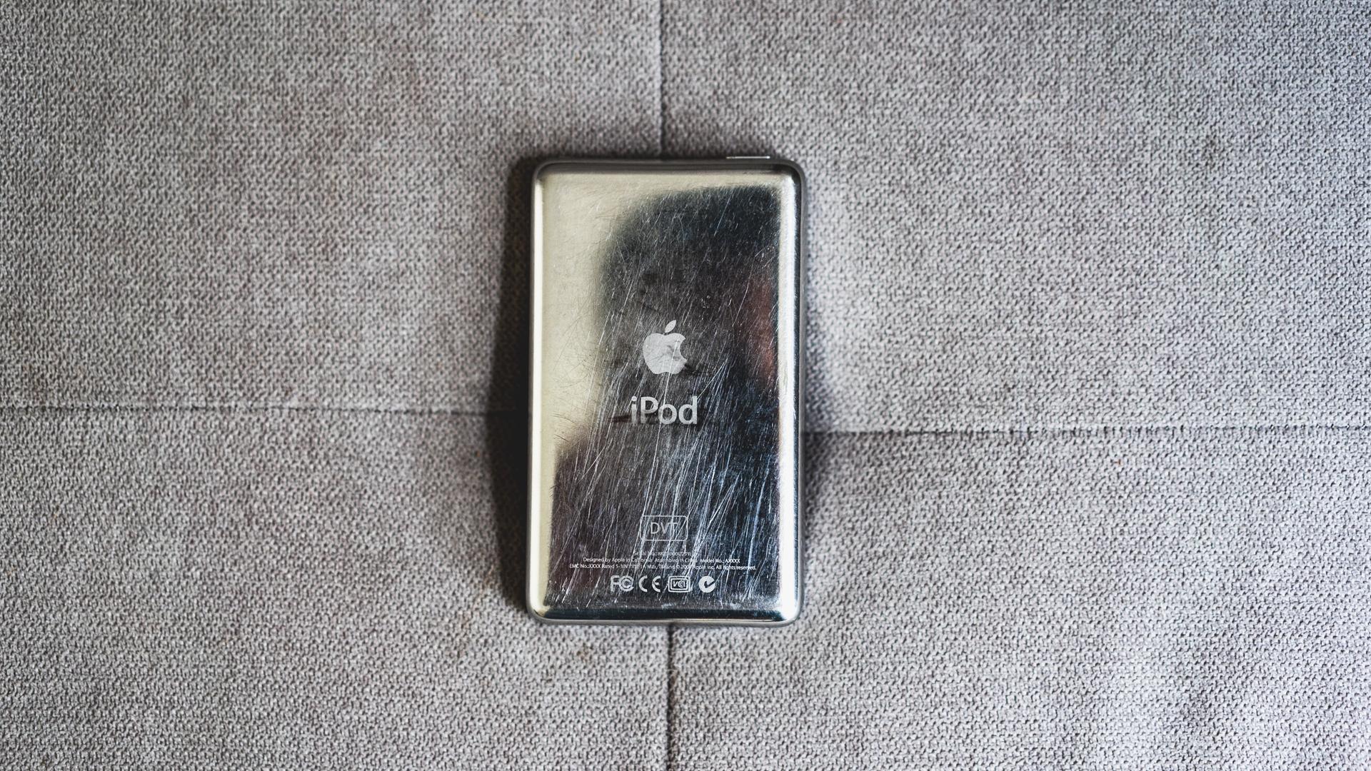 Ein Apple iPod, der Generation 7, liegt auf einer grauen Couch. Zu sehen ist seine, sehr verkratzte und abgenutzte silberne Rückseite. Ein Apple iPod, der Generation 7, liegt auf einer grauen Couch. Zu sehen ist seine, sehr verkratzte und abgenutzte silberne Rückseite.