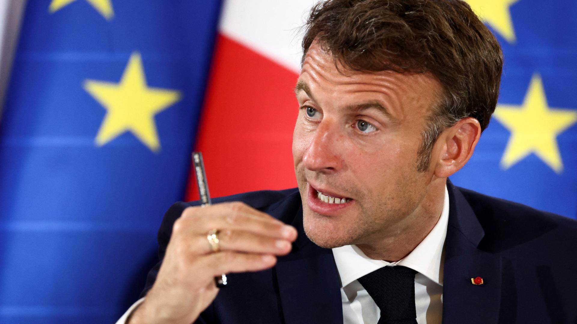 Macron redet engagiert und gestikuliert dabei mit der rechten Hand, in der er einen Stift hält. Dahinter unscharf zwei europäische Flaggen und eine französische.