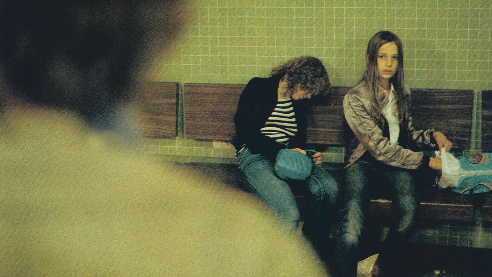 Filmstill aus "Christiane F. - Wir Kinder vom Bahnhof Zoo". Christiane F. sitzt auf einer Bank in einem U-Bahnhof, neben ihr sitzt zusammen gesackt eine weiter Person. Christiane F. schaut zu der Person, die von hinten am linken Bildrand zu sehen ist.