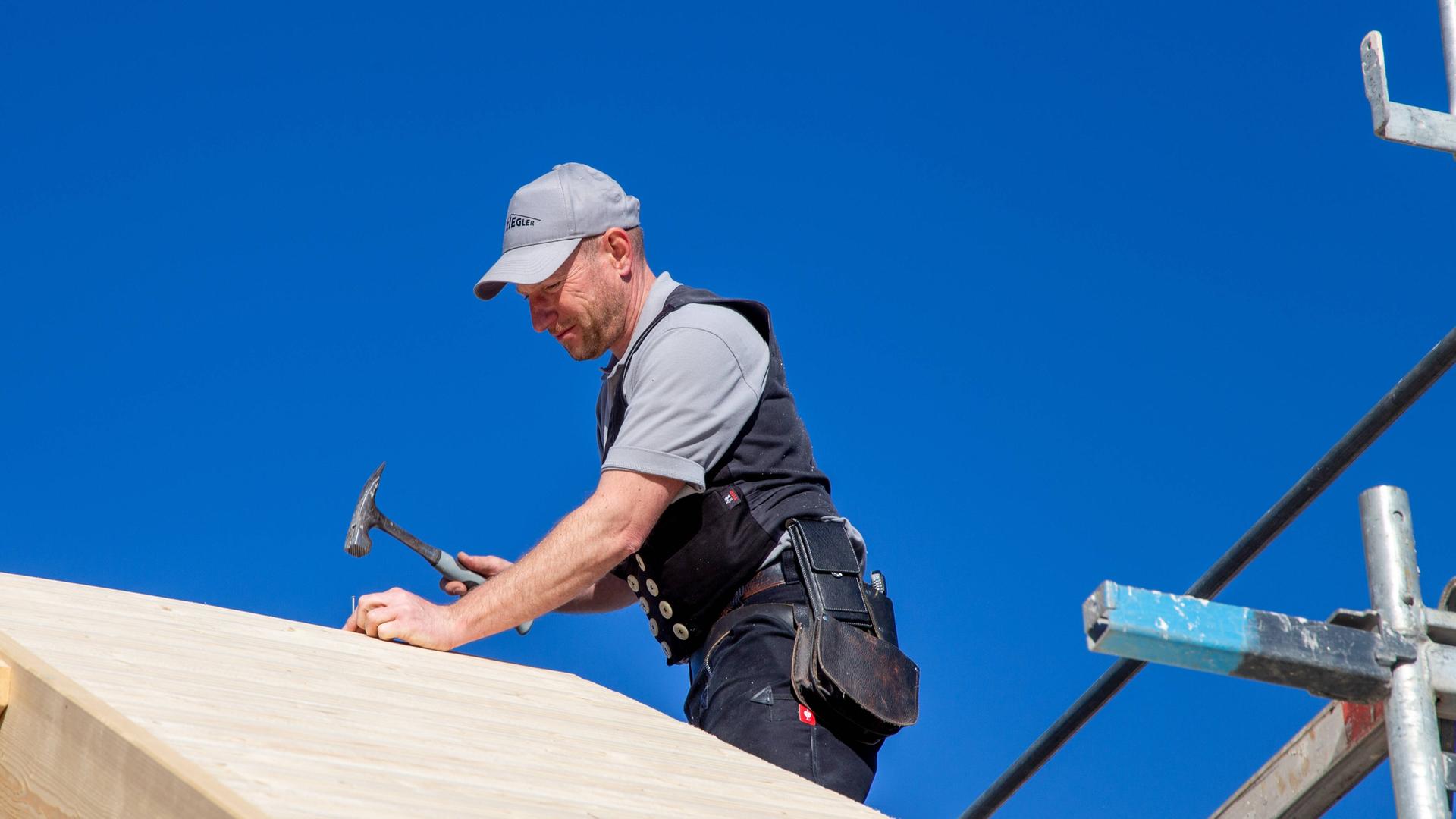 Ein Zimmermann sitzt auf einem Dach und schlägt mit dem Hammer einen Nagel ein.