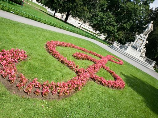 Auf dem Rasen vor dem weißen Mozartdenkmal in Wien sind rosafarbene Blumen angepflanzt, die einen überdimensionalen Notenschlüssel ergeben.