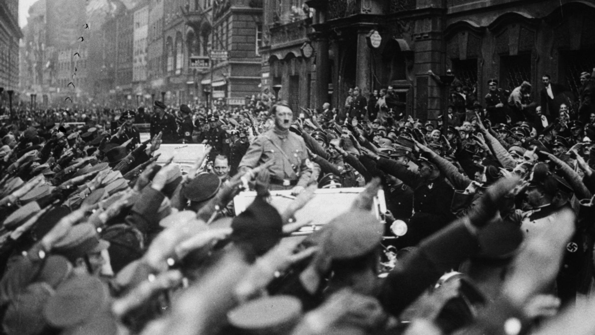 Adolf Hitler fährt stehend auf einem Wagen. An beiden Straßenseiten jubeln ihm Menschen zu. Sie zeigen den Hitlergruß. Adolf Hitler fährt stehend auf einem Wagen. An beiden Straßenseiten jubeln ihm Menschen zu. Sie zeigen den Hitlergruß.