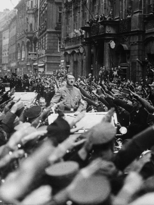 Adolf Hitler fährt stehend auf einem Wagen. An beiden Straßenseiten jubeln ihm Menschen zu. Sie zeigen den Hitlergruß.