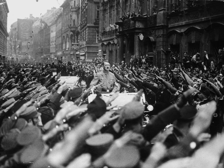 Adolf Hitler fährt stehend auf einem Wagen. An beiden Straßenseiten jubeln ihm Menschen zu. Sie zeigen den Hitlergruß.