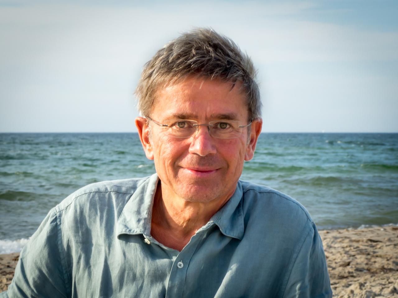 Porträt von Stefan Rahmstorf. Ein mittelalter Mann mit randloser Brille und im blauen Hemd schaut direkt in die Kamera. Hinter ihm ist das Meer zu sehen.
