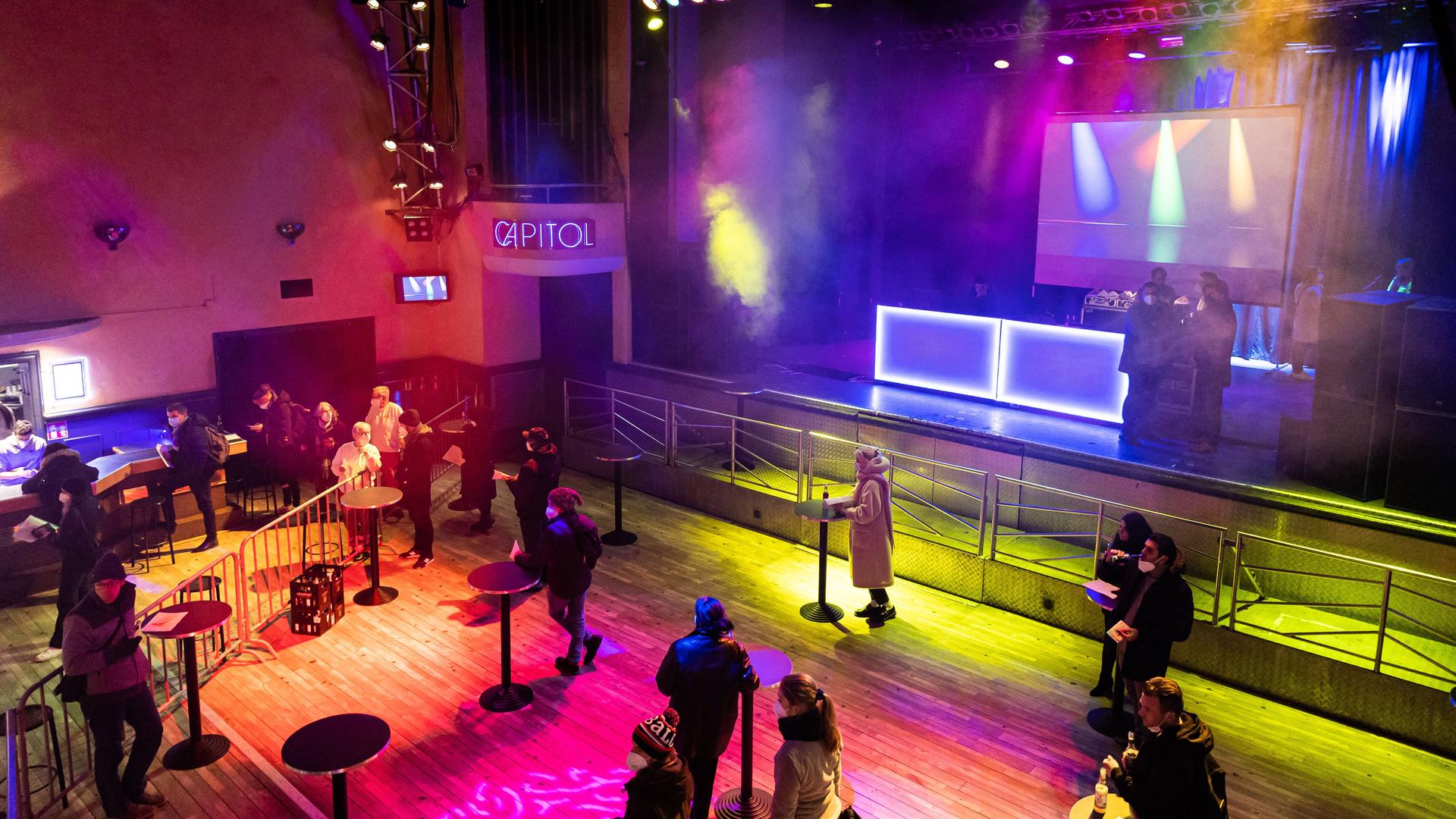 Personen stehen an Stehtischen vor der Bühne im Konzertsaal und warten auf ihre Impfung. Auf der Bühne legt ein DJ Musik auf. Im Konzertsaal des "Capitol" werden vom frühen Abend bis Mitternacht und das gesamte Wochenende Impfungen gegen das Corona-Virus angeboten. Personen stehen an Stehtischen vor der Bühne im Konzertsaal und warten auf ihre Impfung. Auf der Bühne legt ein DJ Musik auf. Im Konzertsaal des "Capitol" werden vom frühen Abend bis Mitternacht und das gesamte Wochenende Impfungen gegen das Corona-Virus angeboten.