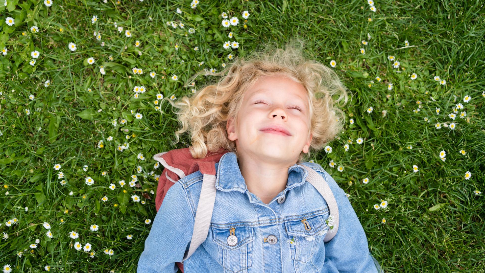 Ein Kind liegt lächelnd mit geschlossenen Augen auf einer grünen Wiese mit Gänseblümchen.