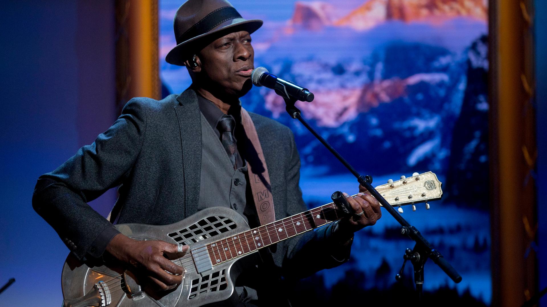 Ein afroamerikanischer Musiker steht mit einer silbernen Gitarre auf der Bühne. Vor ihm steht das Mikrofon. Er trägt einen Hut und einen grauen Anzug. Ein afroamerikanischer Musiker steht mit einer silbernen Gitarre auf der Bühne. Vor ihm steht das Mikrofon. Er trägt einen Hut und einen grauen Anzug.