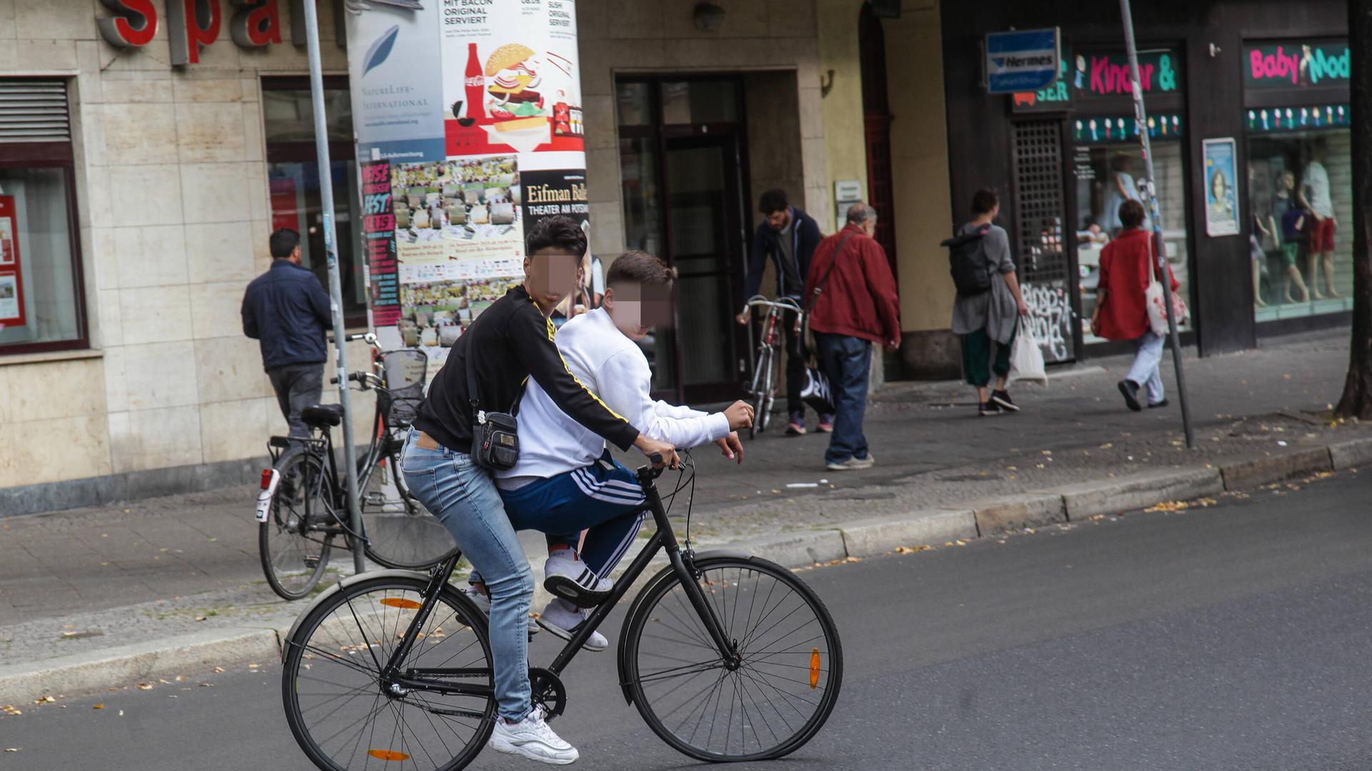 Zwei Jugendliche fahren mit einem Fahrrad – der eine auf der Stange des Fahrrads – auf der Karl-Marx-Allee in Neukölln und behindern den Verkehr. Zwei Jugendliche fahren mit einem Fahrrad – der eine auf der Stange des Fahrrads – auf der Karl-Marx-Allee in Neukölln und behindern den Verkehr.