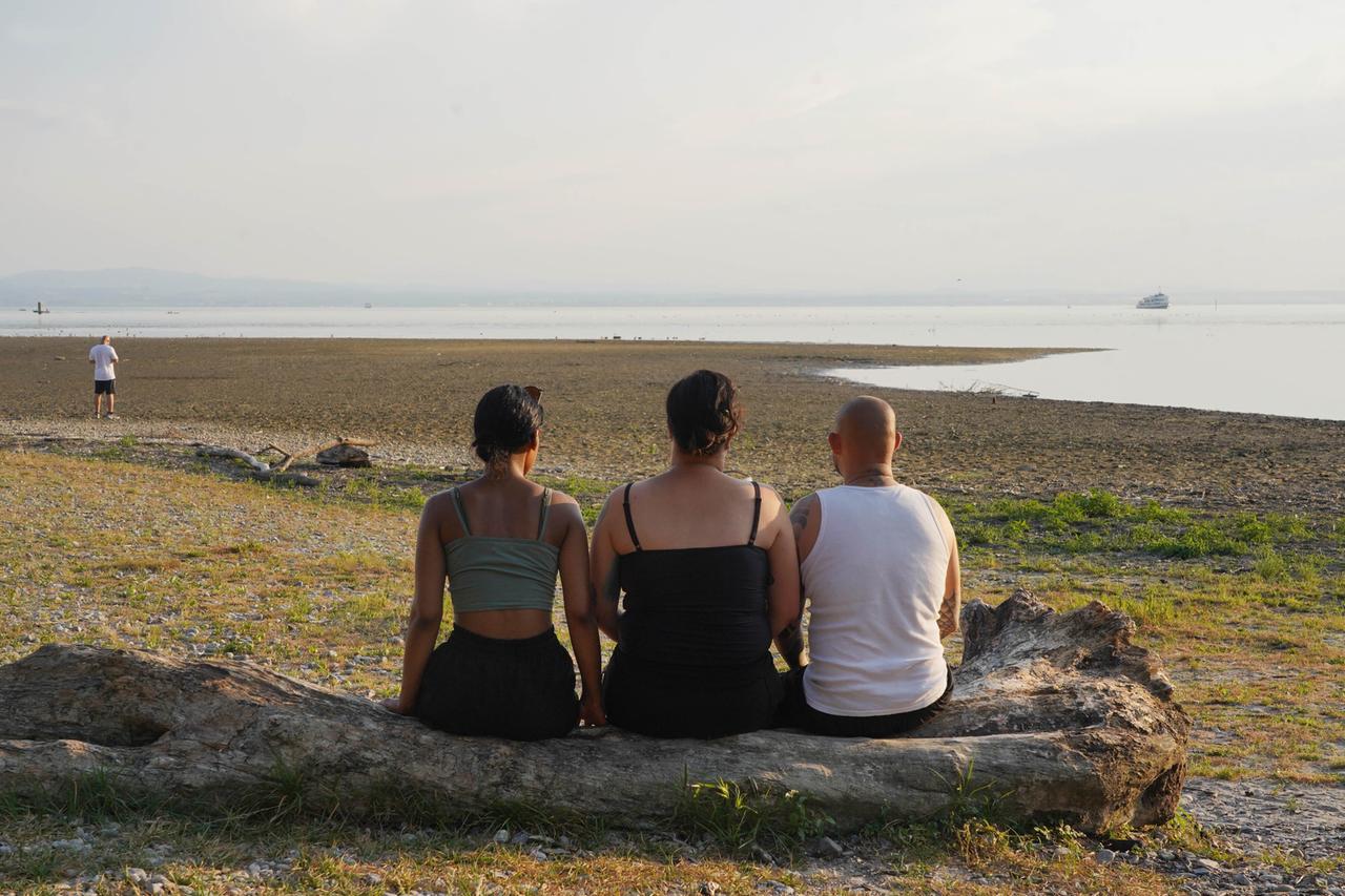 Drei Personen sitzen im versandeten Uferbereich des Bodensees auf einem Baumstumpf.